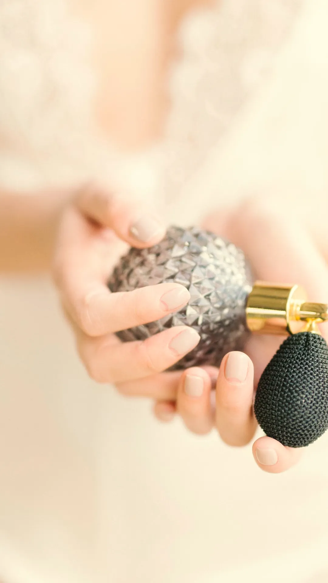 Hands holding a vintage cut-glass bulb atomizer in soft cream light